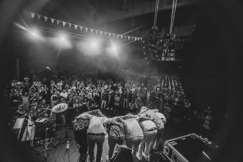 Black and white image of a live concert or event with a large crowd, stage lighting, and performers on stage