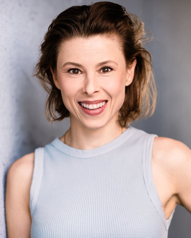 Woman with shoulder-length brown hair smiling at camera wearing light blue sleeveless top against gray background
