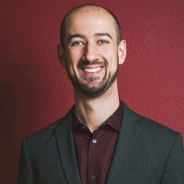 Portrait of a smiling man with short hair and beard wearing a dark blazer and maroon shirt against a red background