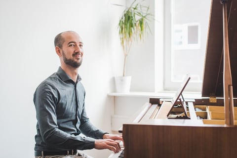 Man in blue shirt playing a grand piano in a bright, modern room with plants and windows